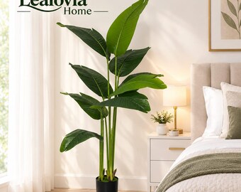 May include: A tall artificial bird of paradise plant with large, vibrant green leaves and sturdy stems, potted in a simple black cylindrical container. The plant features realistic foliage and is positioned upright, displaying a natural leaf structure.