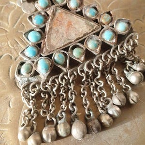May include: A triangular silver-toned pendant featuring a central triangular stone inlay surrounded by small round turquoise-colored cabochons. The piece includes a series of dangling silver-toned chains with small spherical charms at the ends.