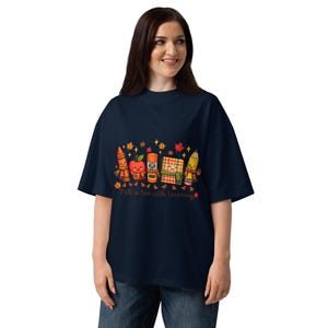 Peut inclure: Un t-shirt bleu marine foncé avec un motif représentant un crayon, une pomme, un bâton de colle, un cahier et un crayon à papier souriants portant des écharpes. Le texte Fall in love with learning est inscrit avec des feuilles d'automne.