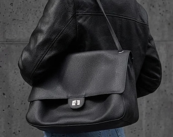 May include: A black leather shoulder bag with a textured surface, a fold-over flap, and a silver-toned metal turn-lock closure. The bag features a thin shoulder strap and is worn over a black leather jacket and blue denim jeans.