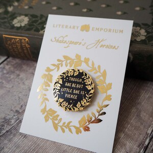 Op de afbeelding: Een zwarte en gouden emaille pin met de tekst "THOUGH SHE BE BUT LITTLE SHE IS FIERCE" op een witte kaart met gouden folie accenten. De kaart leest "Shakespeare's Heroines" en "LITERARY EMPORIUM".