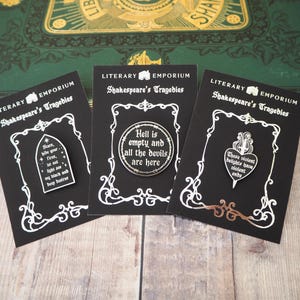 May include: Three black enamel pins featuring Shakespearean quotes on individual black display cards. The pins are shaped like a gothic arch, a circle, and a heart with a dagger, each displaying white text and intricate silver-toned metal detailing.