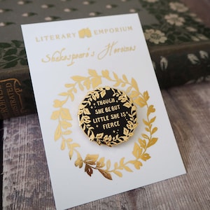 Op de afbeelding: Een goudkleurige pin met de tekst "THOUGH SHE BE BUT LITTLE SHE IS FIERCE" op een zwarte achtergrond, omgeven door een gouden krans. De pin zit op een witte kaart met gouden letters met de tekst "LITERARY EMPORIUM Shakespeare's Heroines".
