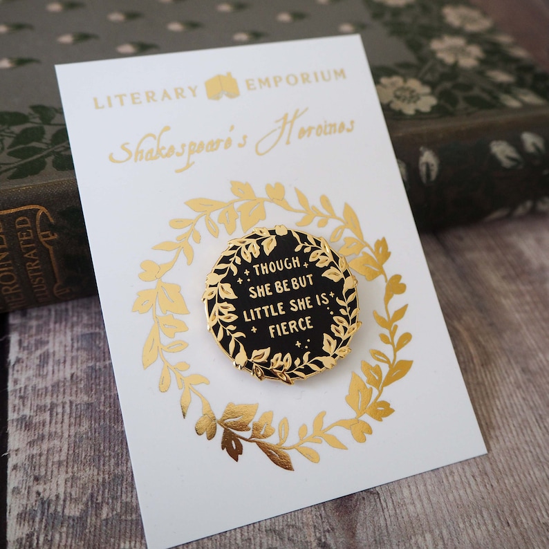 Op de afbeelding: Een goudkleurige pin met de tekst "THOUGH SHE BE BUT LITTLE SHE IS FIERCE" op een zwarte achtergrond, omgeven door een gouden krans. De pin zit op een witte kaart met gouden letters met de tekst "LITERARY EMPORIUM Shakespeare's Heroines".