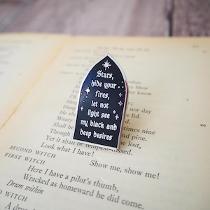 Peut inclure: Une broche en émail noir en forme d'arche gothique, avec un texte argenté qui dit : Stars, hide your fires, let not light see my black and deep desires. Le design comprend de petites étoiles argentées et une bordure en métal argenté.