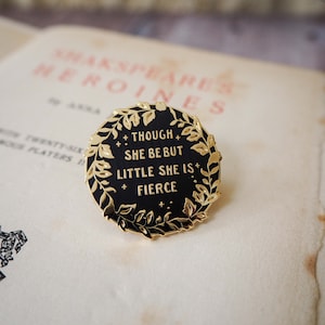 Op de afbeelding: Een zwarte emaille pin met gouden details. De pin toont de quote "Though she be but little she is fierce" omringd door een gouden lauwerkrans. De ronde pin ligt op een vintage pagina van een boek.