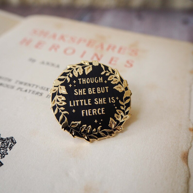 Op de afbeelding: Een zwarte emaille pin met gouden details. De pin toont de quote "Though she be but little she is fierce" omringd door een gouden lauwerkrans. De ronde pin ligt op een vintage pagina van een boek.