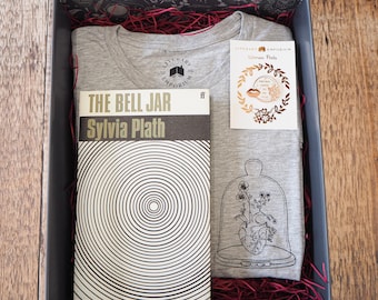 Puede incluir: Una caja de regalo que contiene una camiseta gris con un diseño de corazón floral, una copia de "The Bell Jar" de Sylvia Plath y un pin dorado. La caja está forrada con papel rojo y se encuentra sobre una superficie de madera.
