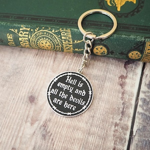May include: A circular metal keyring featuring a black face with white text that reads, "Hell is empty and all the devils are here." The design is bordered by a white barbed wire graphic and includes a silver-toned split ring and chain.