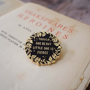 Op de afbeelding: Een ronde, zwarte emaille pin met goudkleurige rand en een laurierkrans ontwerp. De pin bevat de quote "Though she be but little she is fierce" in gouden letters. De pin ligt op een vintage pagina.
