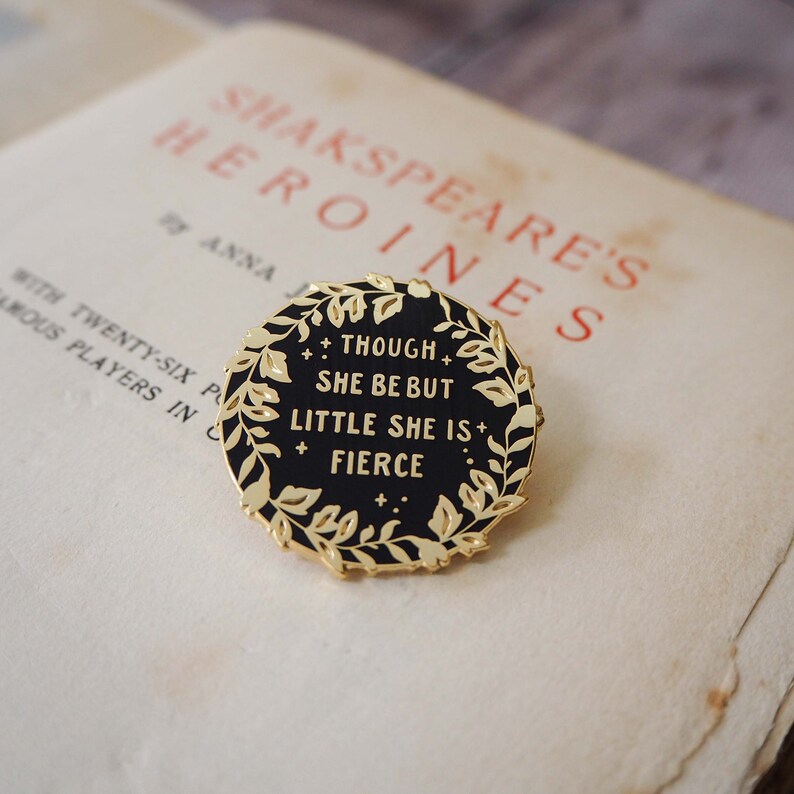 Op de afbeelding: Een ronde, zwarte emaille pin met goudkleurige rand en een laurierkrans ontwerp. De pin bevat de quote "Though she be but little she is fierce" in gouden letters. De pin ligt op een vintage pagina.