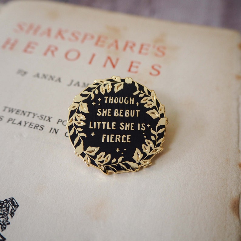 Op de afbeelding: Een ronde zwarte en gouden emaille pin met de quote "Though she be but little she is fierce" omringd door een gouden bladkrans. De pin ligt op een vintage pagina met de titel "Shakespeare's Heroines".