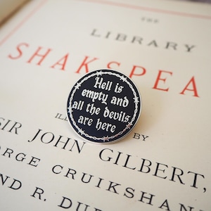 May include: A circular black enamel pin featuring a silver-coloured border of barbed wire. The centre displays the white text "Hell is empty and all the devils are here" written in a gothic-style font, creating a stark contrast.
