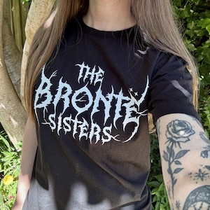 May include: Black crew neck t-shirt with the text "The Bronte Sisters" in a white, gothic-style font. The shirt is made of a dark fabric and is being worn.