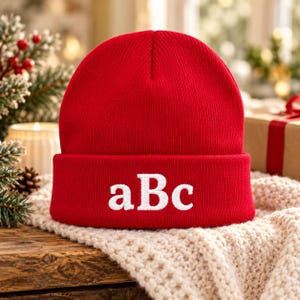 May include: A bright red knit beanie featuring a folded cuff with the letters aBc embroidered in white on the front. The hat is made of a ribbed fabric and has a rounded crown, providing a simple and clean look for cold weather.