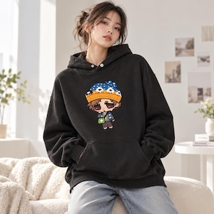 Peut inclure: Un sweat-shirt à capuche noir avec une impression graphique représentant un personnage de dessin animé portant un bonnet bleu et orange à pois blancs, des lunettes de soleil et un sac vert. Il possède des manches longues et une poche kangourou.