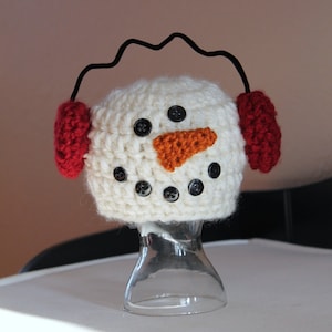 May include: White crocheted snowman hat with red earmuffs and a black button nose. The hat is displayed on a clear glass stand.