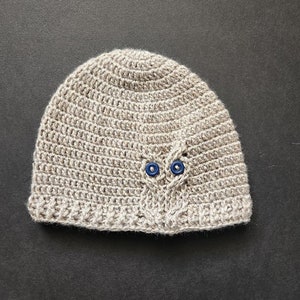 May include: A crocheted beanie hat in a neutral beige color. The hat features a decorative owl design with two blue button eyes. The hat is handmade and suitable for all seasons.