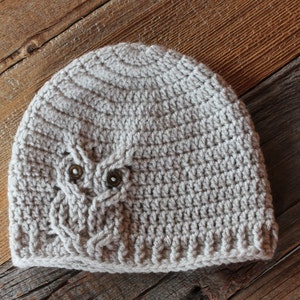 May include: A light gray crocheted beanie hat with an owl design. The owl features two brown button eyes and a textured pattern. The hat is made of a soft, textured yarn and is suitable for all seasons.
