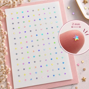 May include: A sheet of small, holographic star-shaped stickers arranged in a grid pattern. A circular inset shows a single star measuring 2 mm in width, placed on a fingertip to demonstrate its scale. The stars reflect various colors.