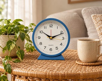May include: A round analog table clock with a blue frame and a white face. The clock features black Arabic numerals, black hour and minute hands, a red second hand, a yellow alarm hand, and the word QUARTZ printed in black on the face.