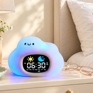 May include: A cloud-shaped digital alarm clock with a glowing blue and white exterior. The front features a black screen displaying the time 06:30, AM, days of the week, a sun icon, a moon icon, and a small battery indicator icon.