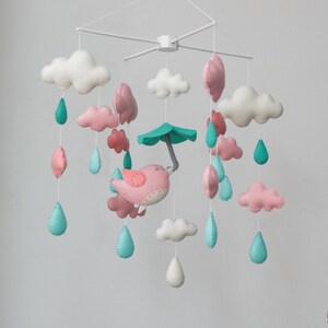 May include: A white baby mobile with pink and white clouds, a pink bird with a green umbrella, and teal raindrops. The mobile is hanging from a white ceiling.