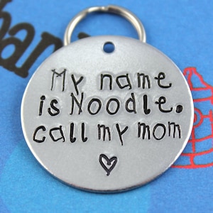 May include: Silver dog tag with a black inscription that reads "My name is Noodle. Call my mom" with a small heart at the bottom.