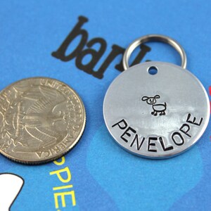 May include: Silver dog tag with a black engraved dog illustration and the name "PENELOPE" on a blue background.