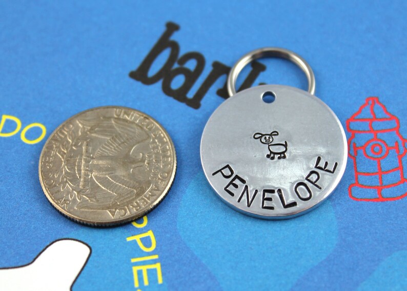 May include: Silver dog tag with a black engraved dog illustration and the name "PENELOPE" on a blue background.