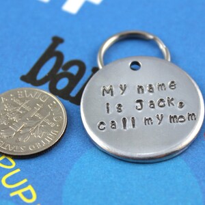Pode incluir: Uma etiqueta de animal de estima&ccedil;&atilde;o de metal prateado com um porta-chaves, estampada com o texto "My name is Jack, call my mom". Uma moeda de dez c&ecirc;ntimos americana est&aacute; ao lado da etiqueta para compara&ccedil;&atilde;o de tamanho. A etiqueta est&aacute; sobre um fundo azul.
