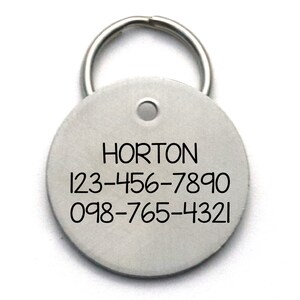 Pu&ograve; includere: Etichetta di identificazione rotonda in argento per animali domestici con un portachiavi. L'etichetta ha il nome "HORTON" e due numeri di telefono: 123-456-7890 e 098-765-4321.