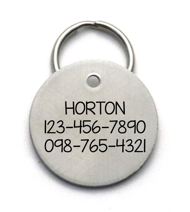 Pu&ograve; includere: Etichetta di identificazione rotonda in argento per animali domestici con un portachiavi. L'etichetta ha il nome "HORTON" e due numeri di telefono: 123-456-7890 e 098-765-4321.