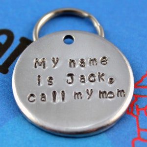 Pode incluir: Uma medalha redonda prateada para animais de estima&ccedil;&atilde;o com um porta-chaves. A medalha tem o texto estampado: "My name is Jack, call my mom."