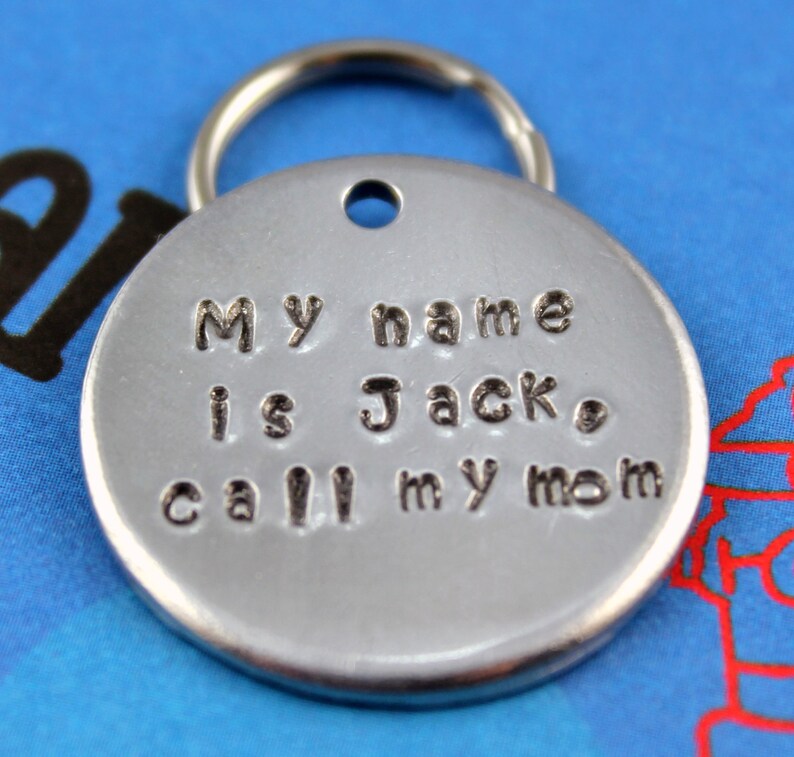 Pode incluir: Uma medalha redonda prateada para animais de estima&ccedil;&atilde;o com um porta-chaves. A medalha tem o texto estampado: "My name is Jack, call my mom."