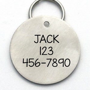 K&ouml;nnte beinhalten: Runder silberner Hundemarkenanh&auml;nger mit schwarzem Ring. Die Marke tr&auml;gt den Namen "JACK" und die Zahlen "123 456-7890" eingraviert.