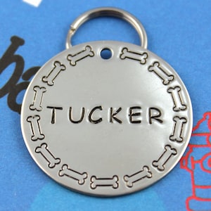 May include: Silver dog tag with a bone design border and the name "TUCKER" engraved in black lettering.
