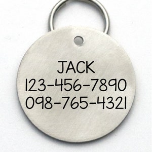 K&ouml;nnte beinhalten: Runder silberner Hundemarkenanh&auml;nger mit einem Ring zum Befestigen am Halsband. Die Marke tr&auml;gt den Namen "JACK" und zwei Telefonnummern: 123-456-7890 und 098-765-4321.
