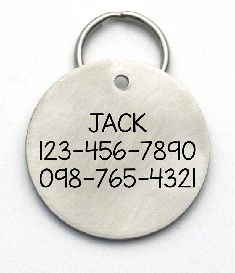 K&ouml;nnte beinhalten: Runder silberner Hundemarkenanh&auml;nger mit einem Ring zum Befestigen am Halsband. Die Marke tr&auml;gt den Namen "JACK" und zwei Telefonnummern: 123-456-7890 und 098-765-4321.