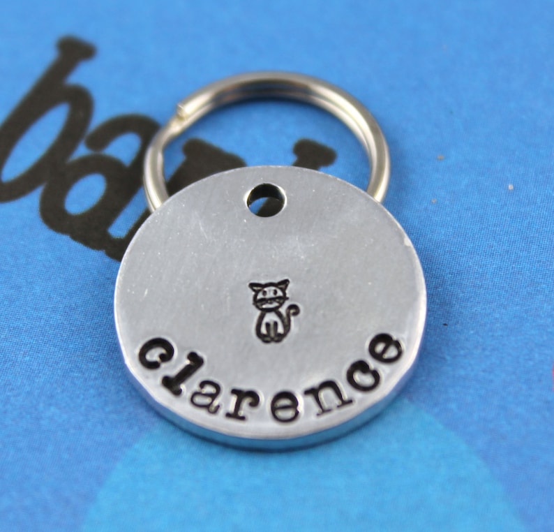 Pu&ograve; includere: Un'etichetta per animali domestici in argento con un'illustrazione di un gatto nero e il nome "Clarence" inciso su di essa.