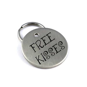 Op de afbeelding: Zilveren sleutelhanger met een rond label waarop "FREE KISSES" in zwarte letters staat.