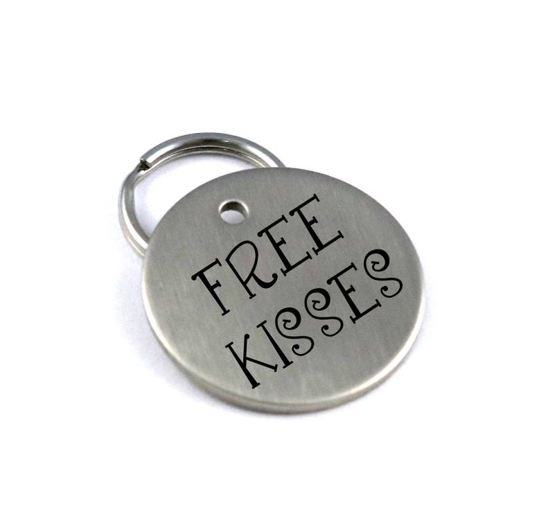 Op de afbeelding: Zilveren sleutelhanger met een rond label waarop "FREE KISSES" in zwarte letters staat.