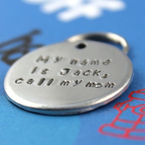 Pode incluir: Uma etiqueta para animais de estima&ccedil;&atilde;o circular prateada com um anel para fixa&ccedil;&atilde;o. A etiqueta tem o texto estampado que diz "My name is Jack, call my mom". O fundo &eacute; azul com uma ilustra&ccedil;&atilde;o de um hidrante.