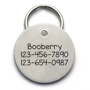 Op de afbeelding: Ronde zilveren hondenpenning met de naam "Booberry" en twee telefoonnummers: 123-456-7890 en 123-654-0987.