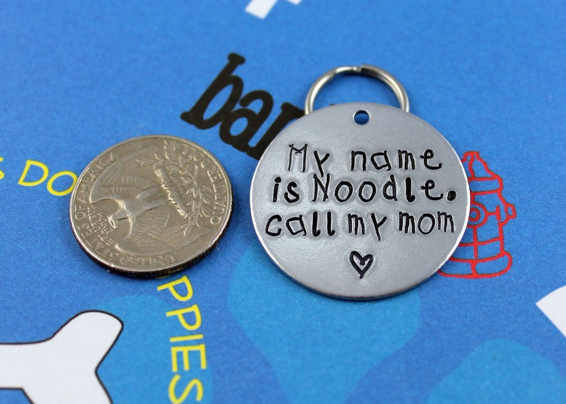 May include: Silver dog tag with a black inscription that reads "My name is Noodle. Call my mom" with a small heart symbol.