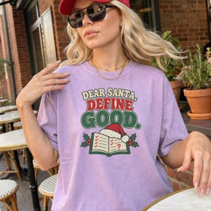 Op de afbeelding: Een lichtpaars T-shirt met ronde hals en een afbeelding met de tekst "DEAR SANTA, DEFINE GOOD." boven een illustratie van een open boek met een rode kerstmuts, groene hulstbladeren en rode bessen op een witte achtergrond.