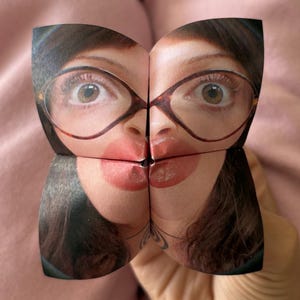 Könnte beinhalten: Ein Papier-Glücksbringer mit einem aufgedruckten Foto eines Gesichts mit braunen Augen, dunklem Haar und einer Brille in Schildpattoptik. Das Bild ist über die vier gefalteten Klappen verteilt und zeigt eine Nahaufnahme von Augen, Nase und Lippen.
