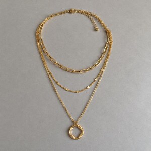 Puede incluir: Un collar de capas en tono dorado con tres cadenas de estilos diferentes. La cadena superior tiene eslabones ovalados gruesos, la central es una cadena de cuentas delicada y la inferior es una cadena fina con un colgante circular trenzado.
