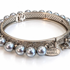 Op de afbeelding: Een scharnierende armband met een rij metallic grijze bolvormige kralen op een zilverkleurige metalen band met filigraanpatroon. Een ovaal metalen label aan de zijkant is gegraveerd met de tekst MIRIAM HASKELL in hoofdletters.