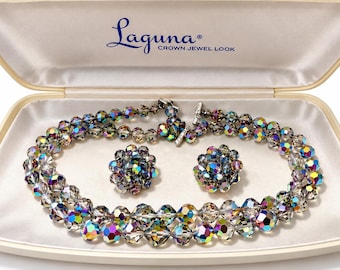Puede incluir: Un conjunto de joyas Laguna Crown Jewel Look que incluye un collar de doble hebra y pendientes de clip a juego hechos de cuentas de cristal facetadas e iridiscentes. El set se presenta en un estuche color crema forrado en satén.